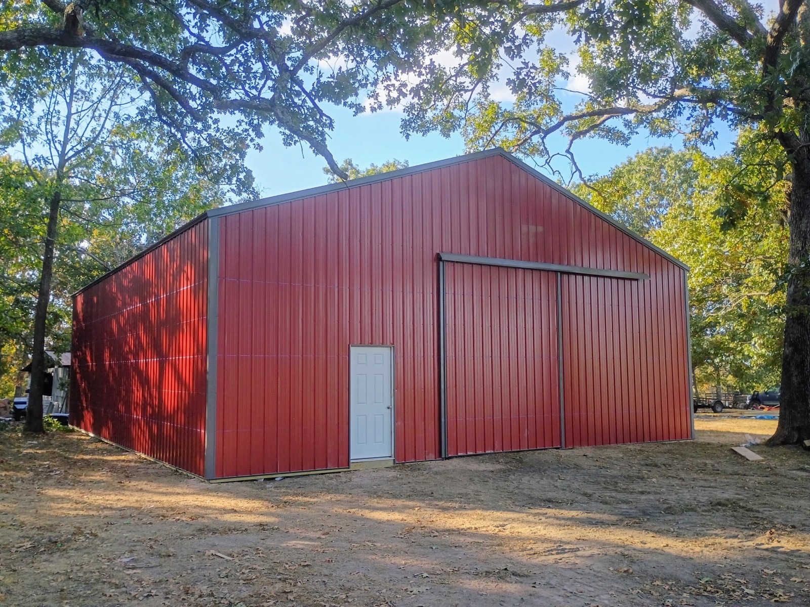 A finished red Lucky pole barn with gray trim, a slider door and a white entry door, under oak trees